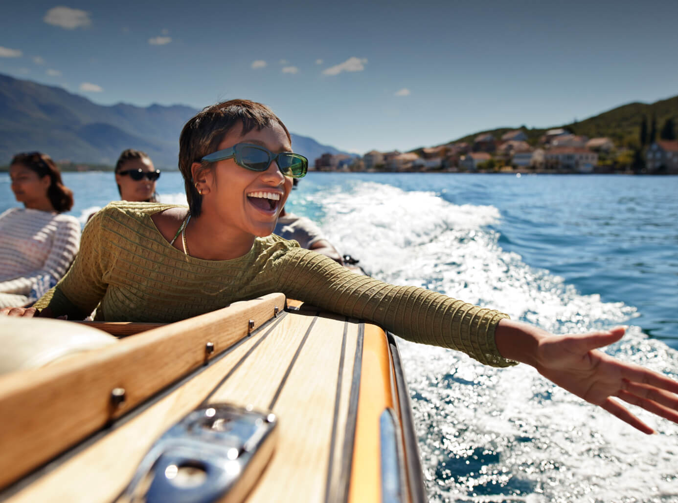 Eine lachende Frau mit Sonnenbrille genießt eine Fahrt auf einem Schnellboot und streckt glücklich eine Hand über das Wasser.