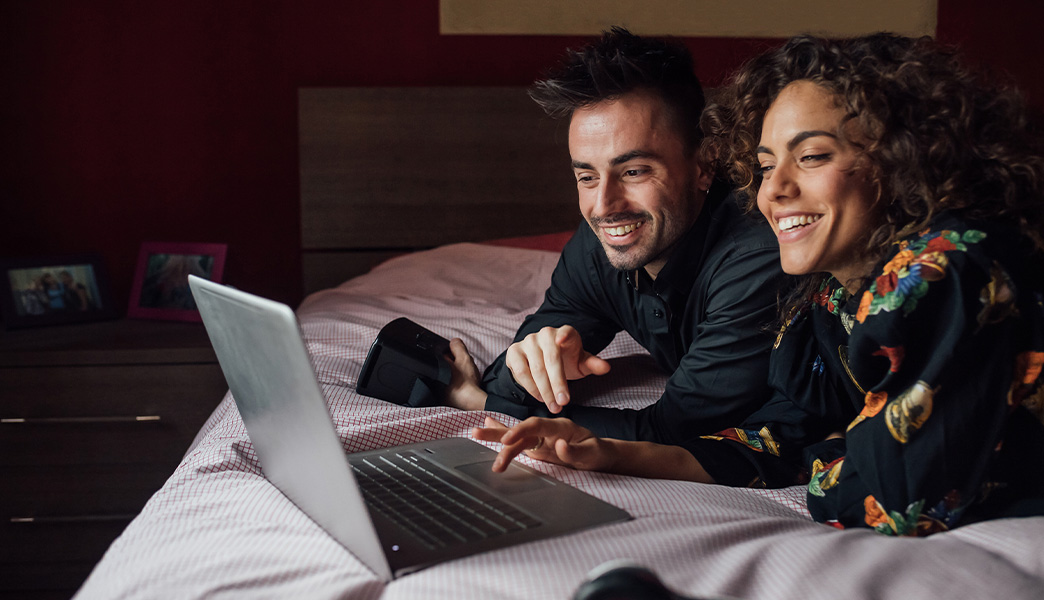 Paar liegt mit einem Laptop auf dem Bett und bucht Tickets 