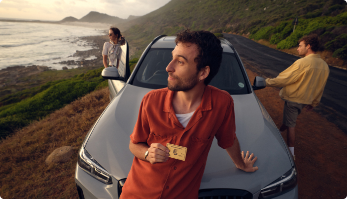 Ein Mann sitzt mit einer Gold Card auf seinem Auto und schaut aufs Meer