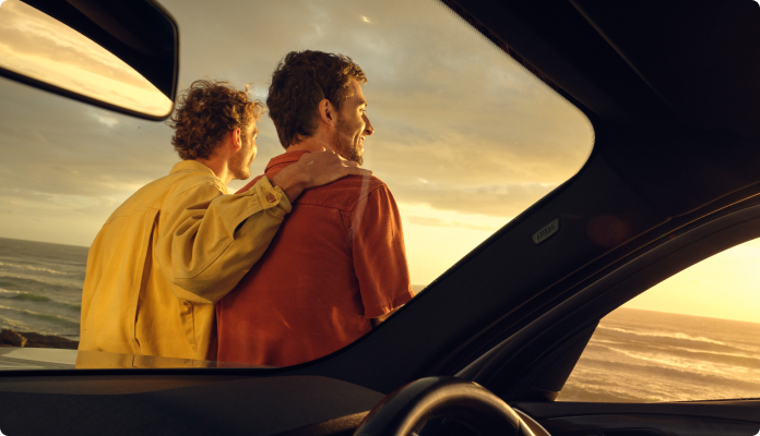 Zwei Freunde sitzen auf der Motorhaube ihres Autos und schauen aufs Meer